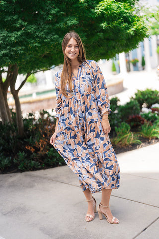 A full-length view of a smiling woman posing outdoors in the Seaside Mosaic Midi Dress—a breezy, bohemian tiered midi dress featuring an artistic abstract brushstroke pattern in slate blue, deep navy, and sandy beige tones. She gently lifts the hem of the relaxed, multi-tiered skirt to highlight its volume and movement, styled with nude ankle-strap block heel sandals.