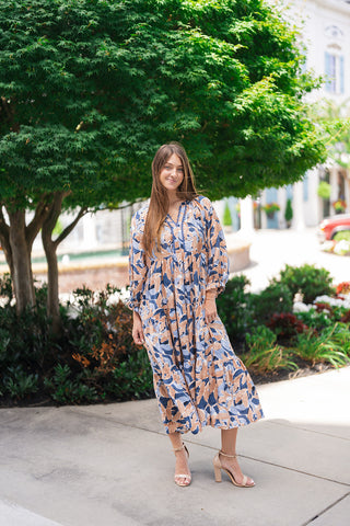 A full-length front view of a model standing in front of lush green trees wearing the Seaside Mosaic Midi Dress. The image shows the modest, comfortable fit of the 3/4 sleeves, the flattering empire waistline, and the flowy, three-tiered midi skirt falling gracefully to mid-calf level above nude heels.