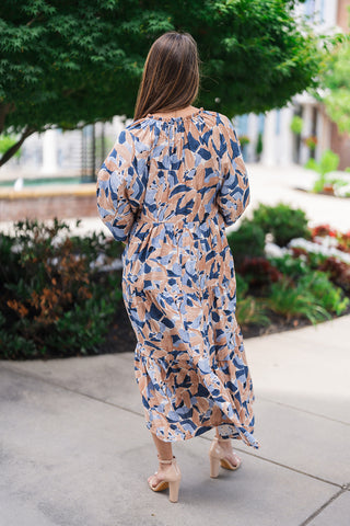 Back view of the Seaside Mosaic Midi Dress demonstrating the uninterrupted flow of the abstract blue and sand print across the gathers and multi-tiered skirt. The shot showcases the gathered neckline detail, full back coverage, and the effortless, airy movement of the hemline as she walks outdoors.
