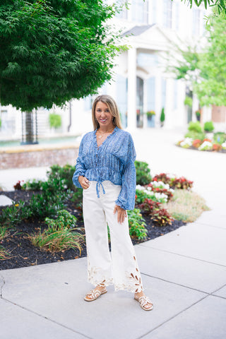 A full-length front view of a woman smiling and posing in the Watercolor Waves Blouse, an ethereal periwinkle blue top featuring a swirling abstract watercolor print. The lightweight, semi-sheer blouse is designed with a smocked waist, front tie accents, an open ruffled V-neckline, and billowy balloon sleeves. She styles the romantic top with high-waisted white wide-leg trousers featuring cutout floral embroidery at the hem, gold layered coin necklaces, and flat tan slides.