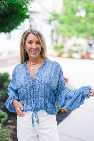 A mid-length front view showcasing the romantic, artisanal silhouette of the Watercolor Waves Blouse. The sheer periwinkle blue woven fabric displays a fluid paisley and abstract watercolor wave design. This portrait shot highlights the open V-neckline outlined by a delicate ruffle trim, button-front closure, and an intricately smocked elasticated waist finished with a front tie accent. The dramatic balloon sleeve is held out to show its lightweight volume.