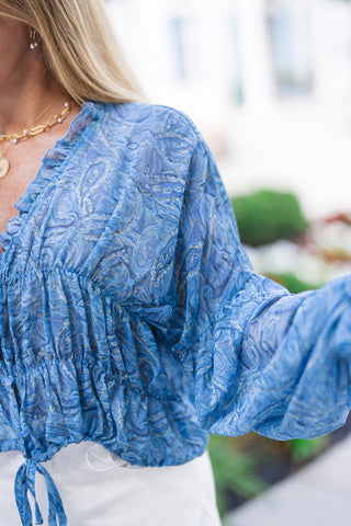 A detailed close-up side angle focusing on the rich texture and construction of the Watercolor Waves Blouse. The image highlights the intricate smocking along the elasticated waist panel and the sheer, crinkled texture of the fluid periwinkle blue fabric. The billowy balloon sleeve extends outward, showing off its generous volume and soft drape.