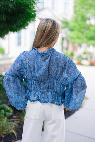 A full-length back view of the Watercolor Waves Blouse, highlighting the uniform gathering from the wide smocked waist panel and a subtle ruffled trim along the high collar. The fluid periwinkle blue printed fabric shows a semi-sheer texture against the light. The blouse is styled with white wide-leg pants, offering a sophisticated, airy ensemble perfect for a resort vacation or a summer evening out.