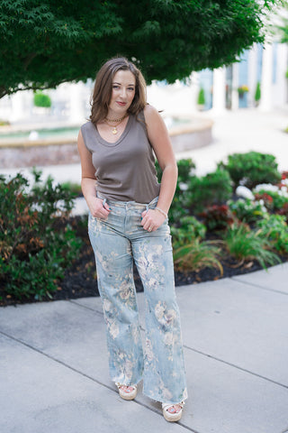 Woman posing outdoors on a concrete sidewalk wearing the Z Supply Ellie V-Neck Tank in a rich taupe-brown shade. The fitted basic tank top is paired with high-waisted, light-wash denim trousers decorated with a faded vintage floral print. Styled with cream-colored platform wedge sandals and gold jewelry against a lush green tree backdrop.