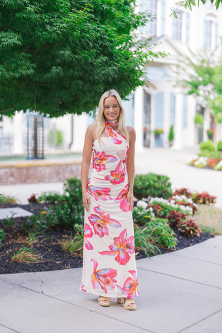 A smiling blonde woman posing outdoors on a sidewalk wearing the Hawaiian Hibiscus One-Shoulder Maxi Dress. The lightweight, ankle-skimming gown features a vibrant oversized floral print in hot pink, coral red, soft orange, and pool blue against a crisp white background. Designed with an asymmetrical halter-style strap, a body-contouring bodice, and dense side-ruching, the look is styled with tan platform block-heel sandals.
