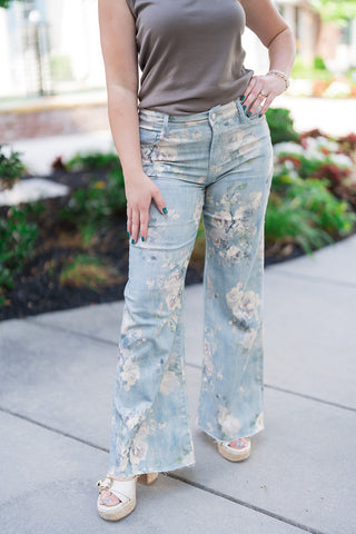 A lower-body front shot of the Dear John Micah Sea Glass Jeans highlighting the relaxed fit that flows into a contemporary clean hem flare silhouette. The view showcases the classic five-pocket construction, secure button-and-zip fly closure, and the painterly floral details, paired with open-toe platform sandals featuring a gold buckle.
