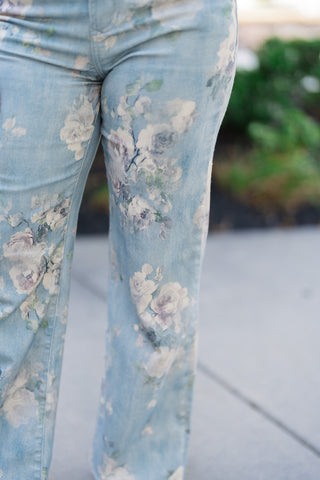 A detailed macro shot focusing on the premium stretch fabric of the Dear John Micah Sea Glass Jeans. The close-up emphasizes the rich texture of the denim and the intricate, weathered details of the faded cream, dusty sage green, and soft lavender blossoms layered over the soft light blue wash.