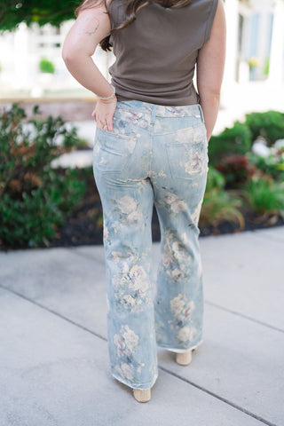 Back view of the Dear John Micah Sea Glass Jeans showcasing the clean fit and traditional rear pocket structuring. The continuous vintage botanical floral print wraps cleanly around the back thighs and waist, highlighting how the mid-rise cut contours the body comfortably, styled with her hand resting in the back pocket.