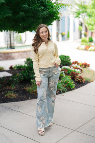 A full-length front view of a woman smiling and posing in the Z Supply Cera Lemon Cardigan, a preppy, slightly cropped cable knit top in a delicate pale lemon yellow hue. The cardigan features a clean crew neckline and a full button-front placket with matching tonal buttons. She has styled the sweater with high-waisted, wide-leg denim jeans covered in a soft blue and cream floral pattern, completed with platform wedge sandals on an outdoor walkway.