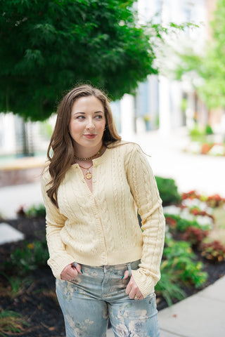 A mid-length front view highlighting the tailored, slim fit of the Z Supply Cera Lemon Cardigan worn partially unbuttoned as a standalone top. The premium cotton-blend knit showcases an allover classic vertical cable yarn stitching pattern running down the body and long sleeves. The look is accessorized with a layered gold link and coin charm necklace, paired with light-wash floral-printed jeans.