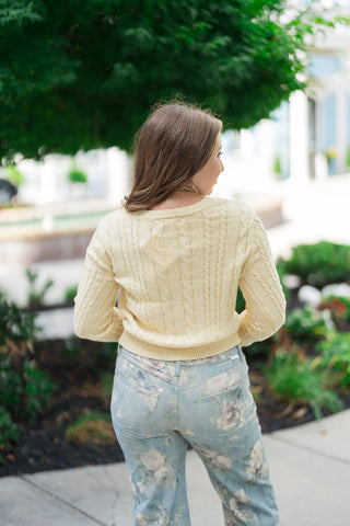 A full-length back view of the Z Supply Cera Lemon Cardigan, showcasing the clean alignment of the vertical cable knit patterns running evenly across the shoulders and back. The slightly cropped hem rests naturally right above the hips of her high-waisted floral denim jeans, emphasizing a sleek, bulk-free profile in an outdoor plaza setting.