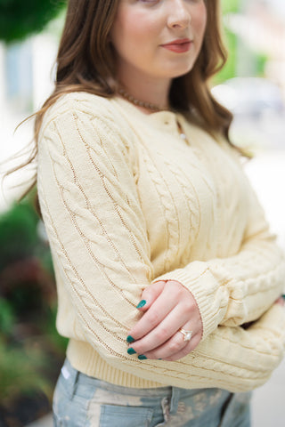 A detailed close-up shot focusing on the sleeve and texture of the Z Supply Cera Lemon Cardigan. The macro view displays the rich visual dimension of the raised vertical cable stitching pattern and fine sweater knit loops. The model has her arms loosely crossed, highlighting the structured ribbing at the cuffs and the soft, breathable quality of the pale yellow fabric.