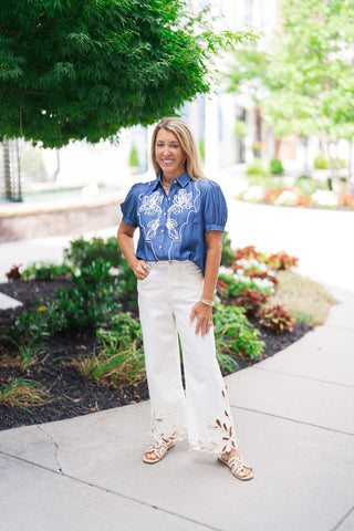 A smiling woman posing outdoors on a concrete sidewalk wearing the Canyon Rosewood Embroidered Top. The lightweight, dark-wash chambray blouse features short puff sleeves and is tucked cleanly into high-waisted wide-leg white jeans that showcase intricate, laser-cut floral details along the hemline. The western-chic look is styled with tan leather slide sandals and minimal gold jewelry against a blurred park background.