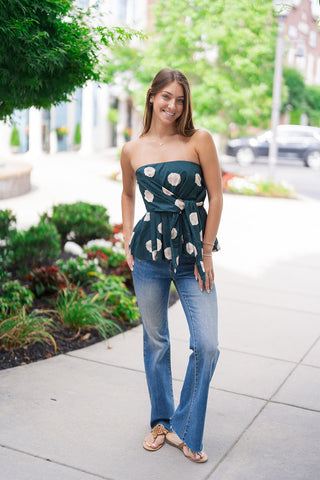A smiling woman posing outdoors on a concrete path wearing the Gallery Stroll Strapless Top. The structured strapless piece features an artistic, large-scale painterly polka dot pattern against a deep forest green backdrop. Styled for an upscale casual daytime look, she pairs the peplum-flared top with high-waisted flared blue denim jeans and flat tan leather thong sandals.