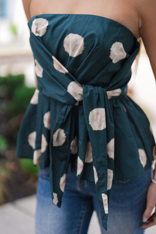 A detailed, waist-level close-up view focusing on the front construction of the Gallery Stroll Strapless Top. The shot highlights the dramatic self-tie feature where fabric panels cross over the bust and secure into a knot at the waist, naturally transitioning into a voluminous peplum flared hemline against blue denim.
