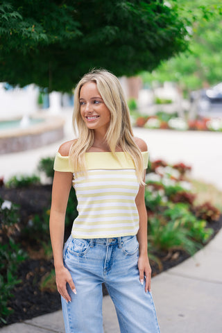 A smiling blonde woman posing outdoors along a sidewalk wearing the Sunny Side Off-The-Shoulder Top. The lightweight, fitted knit top features alternating soft pastel yellow and white horizontal stripes with a prominent, thick fold-over detail framing the neckline. She pairs the retro-chic, sleeveless cropped top with high-waisted light-wash wide-leg denim jeans against a lush green park backdrop.