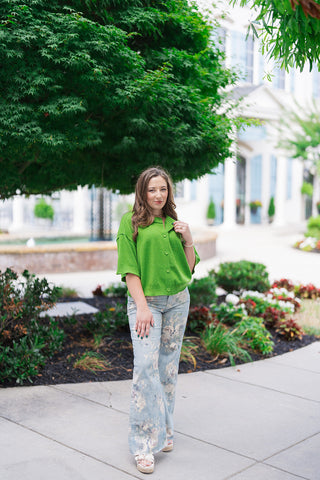 Woman posing outdoors on a sidewalk wearing the Metropolitan Mod Top, a solid chartreuse-lime green short-sleeve button-front knit jacket. The top features a relaxed boxy silhouette with dramatic dropped shoulders and a premium ribbed knit hemline. She pairs the vibrant chartreuse top with high-waisted, light-wash denim trousers adorned with a faded vintage floral print, completing the look with cream platform sandals.