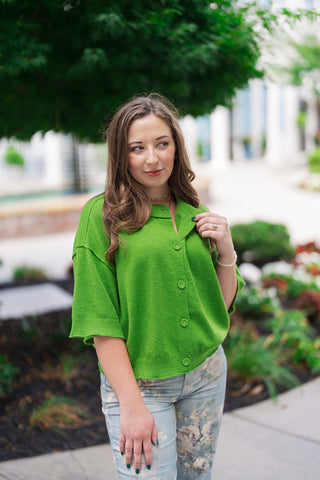 A medium close-up portrait view of the Metropolitan Mod Top highlighting its vintage sixties-inspired mod details. The image showcases the clean round neckline, structured short wide sleeves, and large, color-matched fabric-covered buttons running down the front placket. The chartreuse-lime green woven knit fabric offers a soft, premium texture, beautifully accessorized with a subtle gold chain necklace.