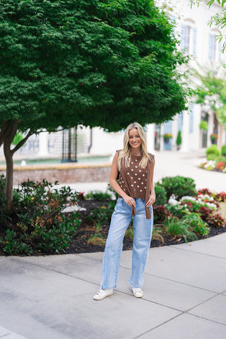 A smiling blonde woman posing outdoors next to a landscaped garden path wearing the Nantucket Knit Dot Top in the Brown colorway. The sleeveless sweater knit tank features an allover cream polka dot motif and a built-in, attached solid brown knit scarf tie detail draped over the shoulders and loosely tied at the front. She pairs the subtly structured, slightly cropped knit top with high-waisted, light-wash blue jeans and white sneakers.
