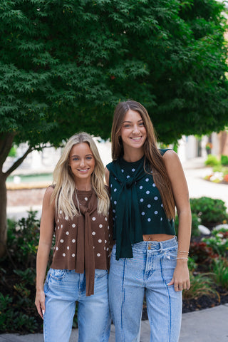 Two women posing side-by-side outdoors under a green tree, showcasing both colorways of the Nantucket Knit Dot Top. The woman on the left wears the Brown and cream variant with light-wash jeans, and the woman on the right wears the Hunter Green and blue variant with seam-detailed denim. The image highlights the versatile neutral options and preppy vintage charm of the sleeveless summer knitwear.