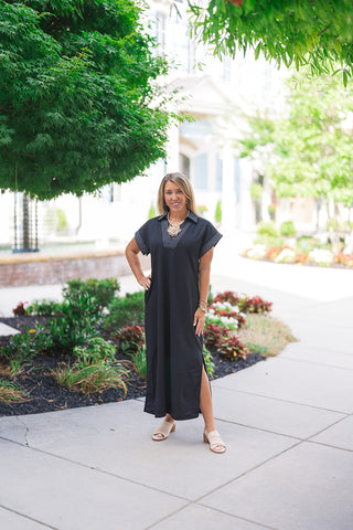 Woman smiling and posing outdoors on a concrete sidewalk wearing the Metropolitan Midi Dress. The solid deep black dress features a relaxed straight silhouette with dynamic side slits along the hemline. Styled for a chic urban summer aesthetic, she pairs the midi dress with cream low block-heeled slide mules and a prominent gold statement necklace in front of a landscaped path.