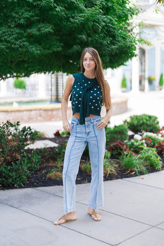 A woman with long brown hair posing outdoors wearing the Nantucket Knit Dot Top in the Hunter Green colorway. The sleeveless tank displays a contrasting royal blue polka dot motif, beautifully accented by the attached green knit scarf tie detail knotted neatly at the crew neckline. She styles the preppy knit top with light-wash denim trousers featuring prominent front center seams and flat tan leather thong sandals.