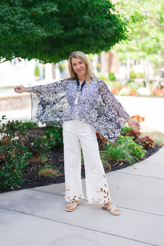 A smiling woman posing outdoors on a concrete sidewalk wearing the Canyon Rosewood Embroidered Top. The lightweight, dark-wash chambray blouse features short puff sleeves and is tucked cleanly into high-waisted wide-leg white jeans that showcase intricate, laser-cut floral details along the hemline. The western-chic look is styled with tan leather slide sandals and minimal gold jewelry against a blurred park background.