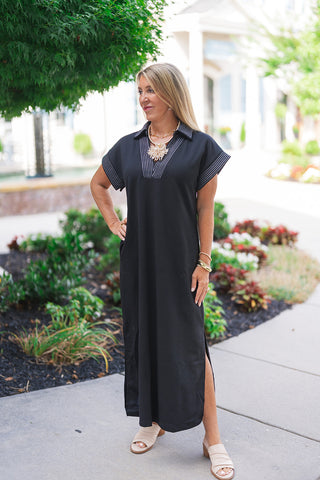A three-quarter full-body view of the Metropolitan Midi Dress highlighting its fluid, unrestrictive drape and functional side slit. The design showcases the contemporary straight cut skimming over the body and discreet built-in side pockets. The model stands with one hand on her hip, accentuating the contrast pinstripe stitching along the V-neckline placket and cap-style sleeves.