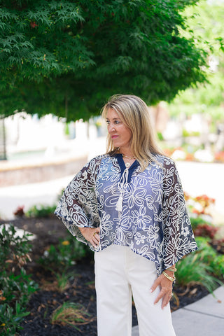 A medium portrait shot of the Canyon Rosewood Embroidered Top highlighting the artistic, dimensional cream-colored rope embroidery that frames the chest and shoulders. The fluid chambray fabric offers the sleek appearance of dark denim, accented by a structured pointed collar, natural wooden-style buttons along the center placket, and romantic gathered puff sleeves. The model completes the look with layered gold chain necklaces and matching bracelets.