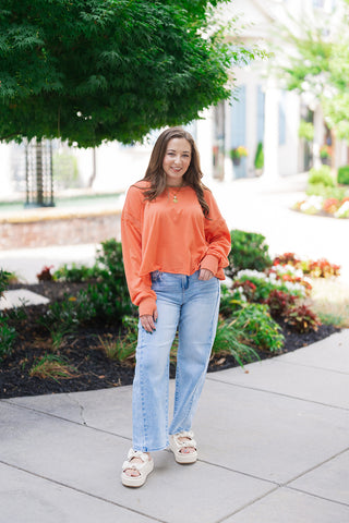 Woman posing outdoors on a concrete sidewalk wearing the Citrus Core Crossover Sweatshirt in a vibrant, sun-drenched orange hue. The relaxed-fit pullover features dropped shoulders and a contemporary raw-edge hemline, styled effortlessly for an athleisure look with light-wash, wide-leg denim jeans and cream-colored platform sandals with bow accents.
