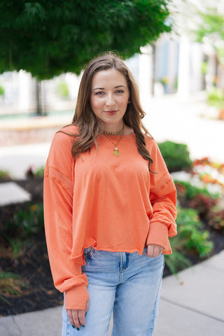 A medium portrait view of the Citrus Core Crossover Sweatshirt showcasing its classic crew neck collar and vibrant citrus orange colorway. The slouchy, comfortable drape of the dropped shoulders is accentuated by a statement layered gold coin pendant necklace, presenting a stylish, off-duty weekend aesthetic.