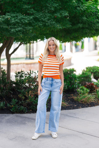 A smiling blonde woman posing outdoors on a concrete sidewalk wearing the Creamsicle Pop Ribbed Top. This short-sleeve knit sweater top showcases a vibrant orange and crisp white horizontal stripe pattern with a detailed vertical ribbed texture. She pairs the retro-inspired top with high-waisted, light-wash wide-leg jeans and clean white sneakers against a backdrop of lush green trees.