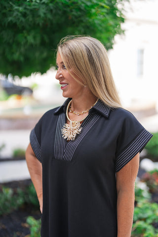 A portrait view of the Metropolitan Midi Dress highlighting the graphic detail of the intricate contrast pinstripe stitching. The crisp white stitched lines define the sharp pointed collar, plunging V-neckline placket, and cap sleeves against the solid black premium knit fabric. The look is accessorized with an ornate, organic-shaped gold statement pendant and a stacked link necklace.