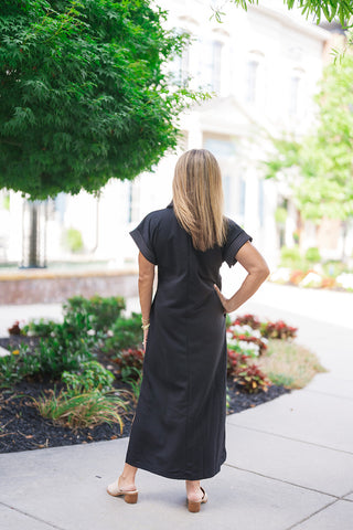 Back view of the Metropolitan Midi Dress showcasing the clean, uninterrupted silhouette and straight-cut profile of the black lightweight knit fabric. This view emphasizes the modern cap-style short sleeves bordered by white contrast stitching lines and illustrates the graceful, modest midi-length coverage from behind as the model poses with one hand on her hip.