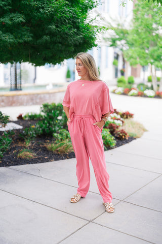 Woman posing outdoors on a sidewalk wearing the Raspberry Sunrise Top styled as a cohesive monochromatic lounge set with the matching relaxed pants. The boxy tee displays a unique, vintage-inspired mineral-washed raspberry pink finish on soft cotton fabric. She stands with her hands in her pockets, completing the effortless look with a gold pendant necklace, stacked gold bracelets, and cream slide sandals.
