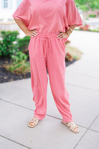 A detailed front close-up view focusing on the waist and leg construction of the Raspberry Sunrise Pants. The image highlights the flexible elastic waistband detailed with a matching functional front drawstring tie. The relaxed-fit legs taper slightly at the bottom, showcasing the unique mineral-wash finish and practical side pockets, paired with cream flat slide sandals.