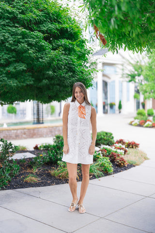 A smiling woman posing outdoors on a concrete sidewalk wearing the Mimosa Morning Eyelet Mini Dress. This crisp white sleeveless shirt dress showcases premium all-over floral eyelet embroidery, a tailored pointed collar, and an adjustable drawstring waist defining its fluid A-line mini skirt. Styled with an orange and white patterned neck scarf and flat white leather thong sandals against a backdrop of lush green trees.