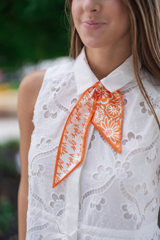A portrait view of the Mimosa Morning Eyelet Mini Dress highlighting the intricate workmanship of the allover floral eyelet cutouts and the crisp structure of the turn-down pointed collar. The close-up showcases a vibrant orange and white patterned silk scarf tied neatly under the collar, adding a pop of color over the dress's hidden button placket.