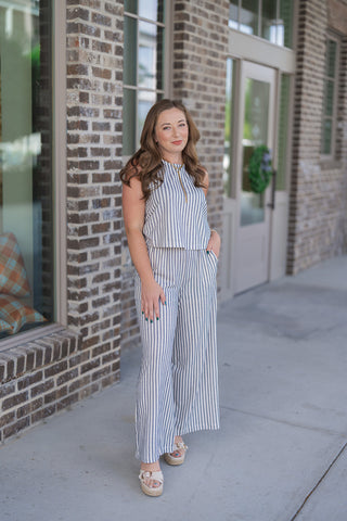 Woman posing outdoors in front of a modern brick storefront wearing the Z Supply Scout Eclipse Striped Pants paired with the matching sleeveless knit top. This complete resort-ready set highlights a cohesive maritime look featuring cream and navy blue vertical stripes. The model stands with one hand tucked into the side pocket, styled with cream-colored platform slide sandals and a bold gold chain necklace.