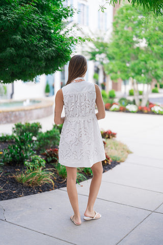 Back view of the Mimosa Morning Eyelet Mini Dress highlighting the continuous, romantic floral eyelet pattern across the entire garment. This view emphasizes the sleeveless profile, the flexible gathered elastic waistline that structures the silhouette, and the thick, solid white band detailing that cleanly outlines the hem of the mini-length skirt.