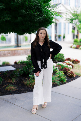 A smiling woman posing outdoors on a sidewalk wearing the Midnight Muse Ruffle Blouse. The lightweight, semi-sheer chiffon top features cascading multi-layered front ruffles along a deep V-neckline and long blouson sleeves with flared bell cuffs. She pairs the romantic black blouse with high-waisted white wide-leg trousers, cream-colored platform heeled sandals, and a gold statement pendant necklace against a lush green park backdrop.