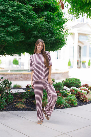 A smiling woman posing outdoors on a concrete path wearing the Wildberry Boardwalk Pants. These lightweight lounge pants feature a beautiful seasonal mauve and dust pink horizontal stripe pattern within a sweeping, flowy wide-leg silhouette. She styles the look as an effortless boutique matching set with the coordinating short-sleeve top and casual brown-and-white sneakers against a lush green tree backdrop.