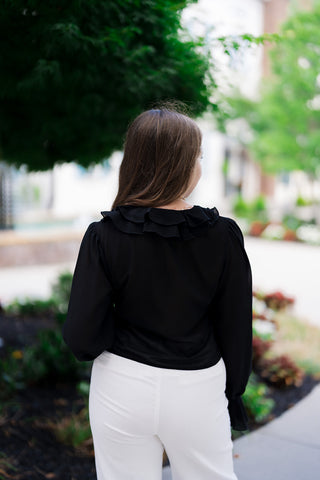 Back view of the Midnight Muse Ruffle Blouse showcasing the clean silhouette and airy drape of the lightweight chiffon fabric from behind. This angle highlights the multi-layered ruffle trim extending across the back of the neck like a sophisticated collar frame. The black top is neatly tucked into the waistband of high-rise white tailored trousers.