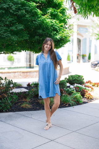 A smiling woman posing outdoors on a sidewalk wearing the Sweet Magnolia Chambray Dress. The soft-wash blue denim mini dress features a relaxed A-line shift silhouette, a clean split V-neckline, and fluid flutter sleeves. She styles the look with flat white leather thong sandals featuring a gold logo medallion against a lush green garden backdrop.