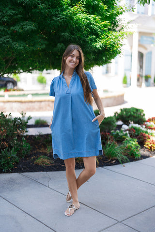 A full-length view of the Sweet Magnolia Chambray Dress showcasing its functional and casual design. The model smiles at the camera with her hands tucked into deep, hidden side-seam pockets. This angle highlights the curved bottom hemline and subtle side slits that enhance the dress's breezy movement.