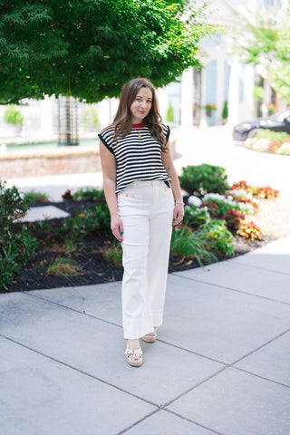 Woman posing outdoors in the Midnight in Paris Sweater Vest, a retro-chic sleeveless knit top featuring a bold black and cream horizontal stripe motif. The relaxed, slightly boxy profile is styled tucked slightly into high-waisted, wide-leg white trousers featuring gold hardware details. She completes the look with open-toe platform wedge sandals and a statement gold pendant necklace.