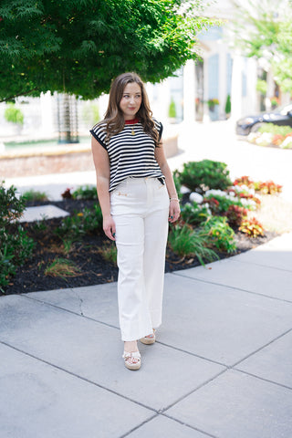 Woman posing outdoors in the Midnight in Paris Sweater Vest, a retro-chic sleeveless knit top featuring a bold black and cream horizontal stripe motif. The relaxed, slightly boxy profile is styled tucked slightly into high-waisted, wide-leg white trousers featuring gold hardware details. She completes the look with open-toe platform wedge sandals and a statement gold pendant necklace.