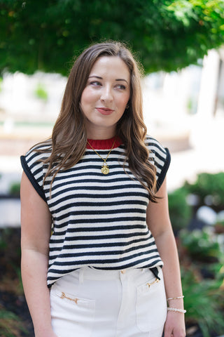 A close-up portrait view of the Midnight in Paris Sweater Vest highlighting its textured knit fabrication and vibrant crimson red ribbed crew collar. The cap-sleeve style armholes are framed with a solid black ribbed trim. Accessorized with a gold coin pendant necklace, the look emphasizes a classic European flair and preppy, elevated summer-into-fall style.