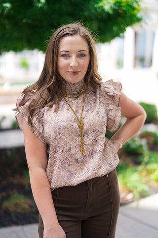 A portrait view of the Serengeti Smocked Mock Neck highlighting the delicate, tailored structure of the elasticized high smocked collar. The close-up showcases the premium woven fabric's modern abstract pattern and the playful, dimensional volume of the double-layered ruffle cap sleeves, accessorized beautifully with a statement gold knot necklace.