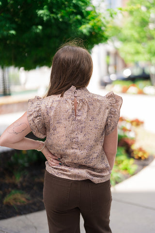 Back view of the Serengeti Smocked Mock Neck showcasing the refined, boutique-quality back neck button and loop closure with a subtle keyhole cutout at the top of the smocked collar. The relaxed-fit earth-toned blouse drapes smoothly over the waistline of dark brown trousers, highlighting the continuous abstract texture and architectural frame of the ruffle sleeves from behind.